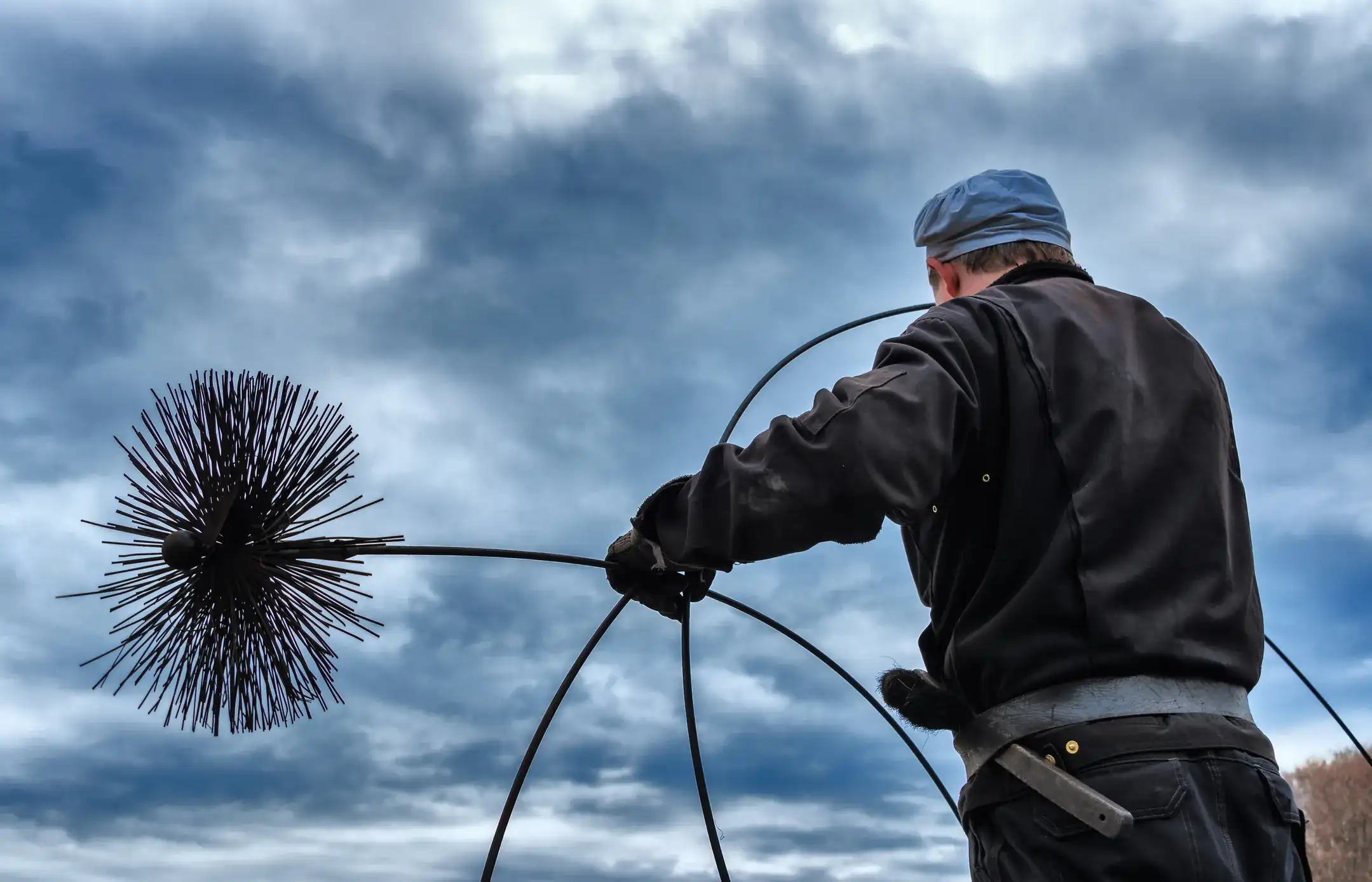 Chimney sweep holding a large round brush on a flexible rod, preparing to clean a chimney flue.
