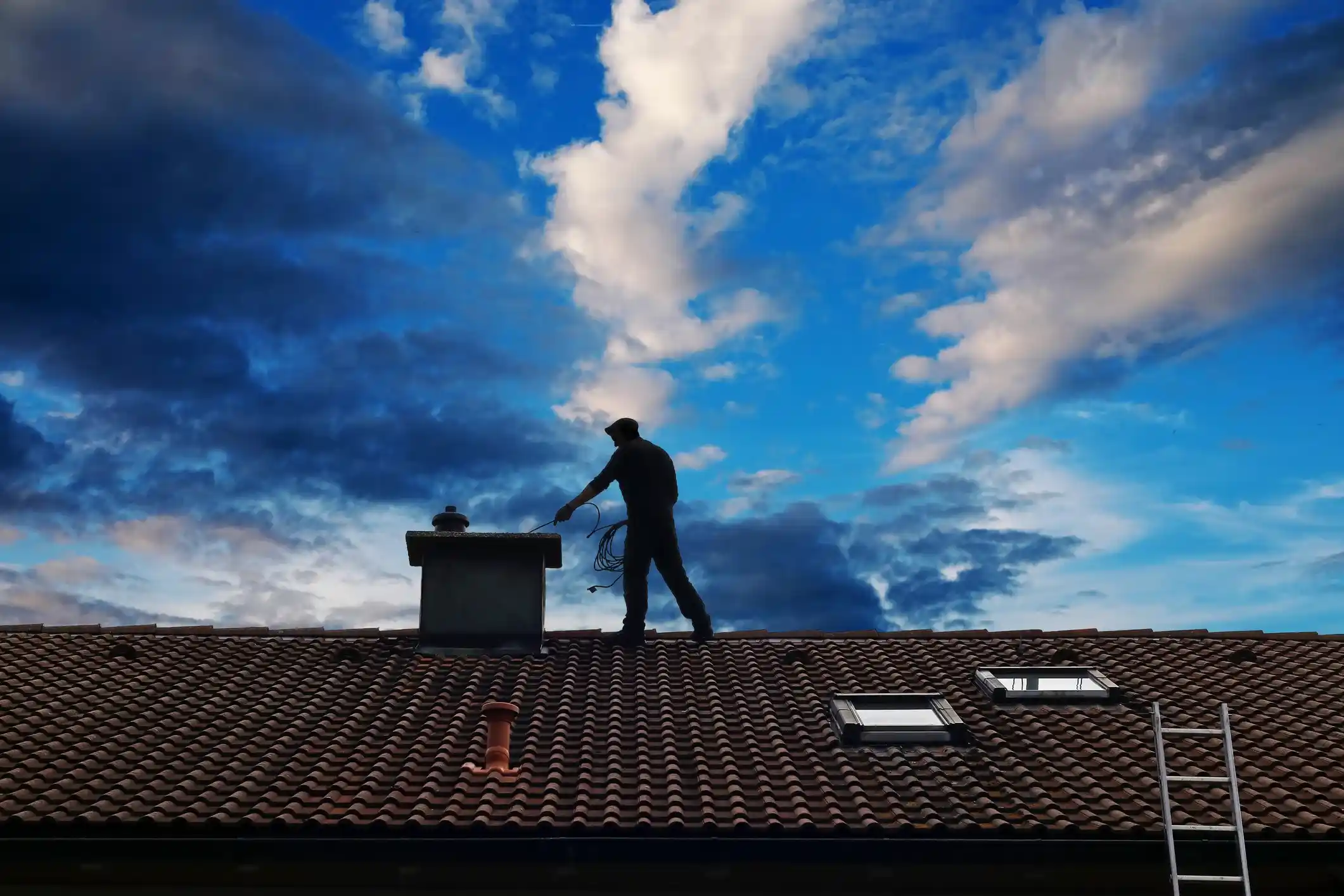 Silhouette of a chimney sweep working on a tiled roof under a dramatically blue, cloudy sky.