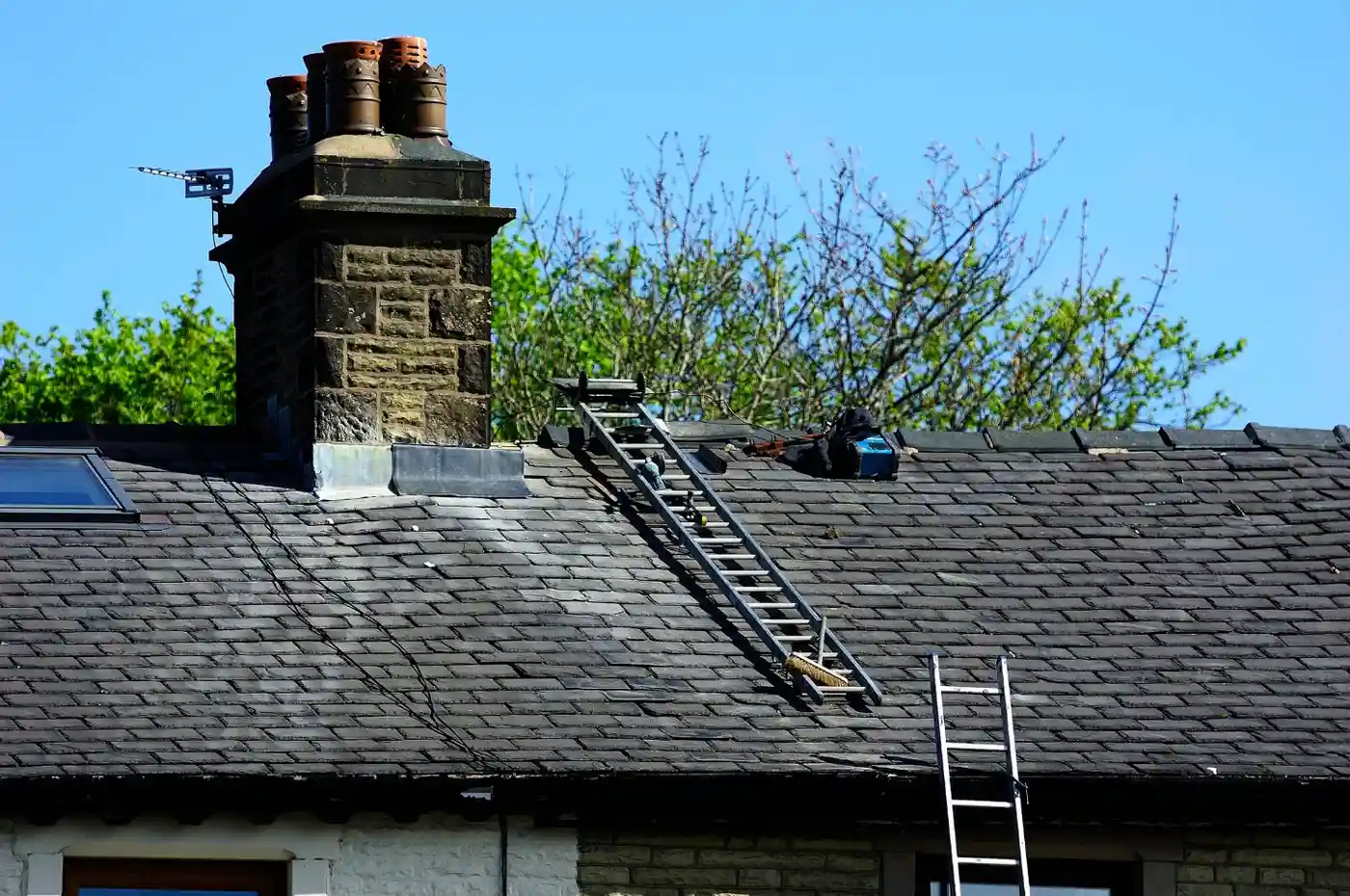 the slate roof of a house on a clear, sunny day, with a focus on the chimney and necessary access equipment. A prominent stone or brick chimney stack with a decorative cap and three terracotta-colored chimney pots stands on the left. A silver, aluminum cat ladder is laid flat on the steeply pitched roof tiles, running up toward the chimney. A dark blue or black tool bag rests near the top of the cat ladder. The lower edge of a standard extension ladder is visible in the bottom right, leaning against the roof. Lush green trees are seen against the clear blue sky above the roofline.