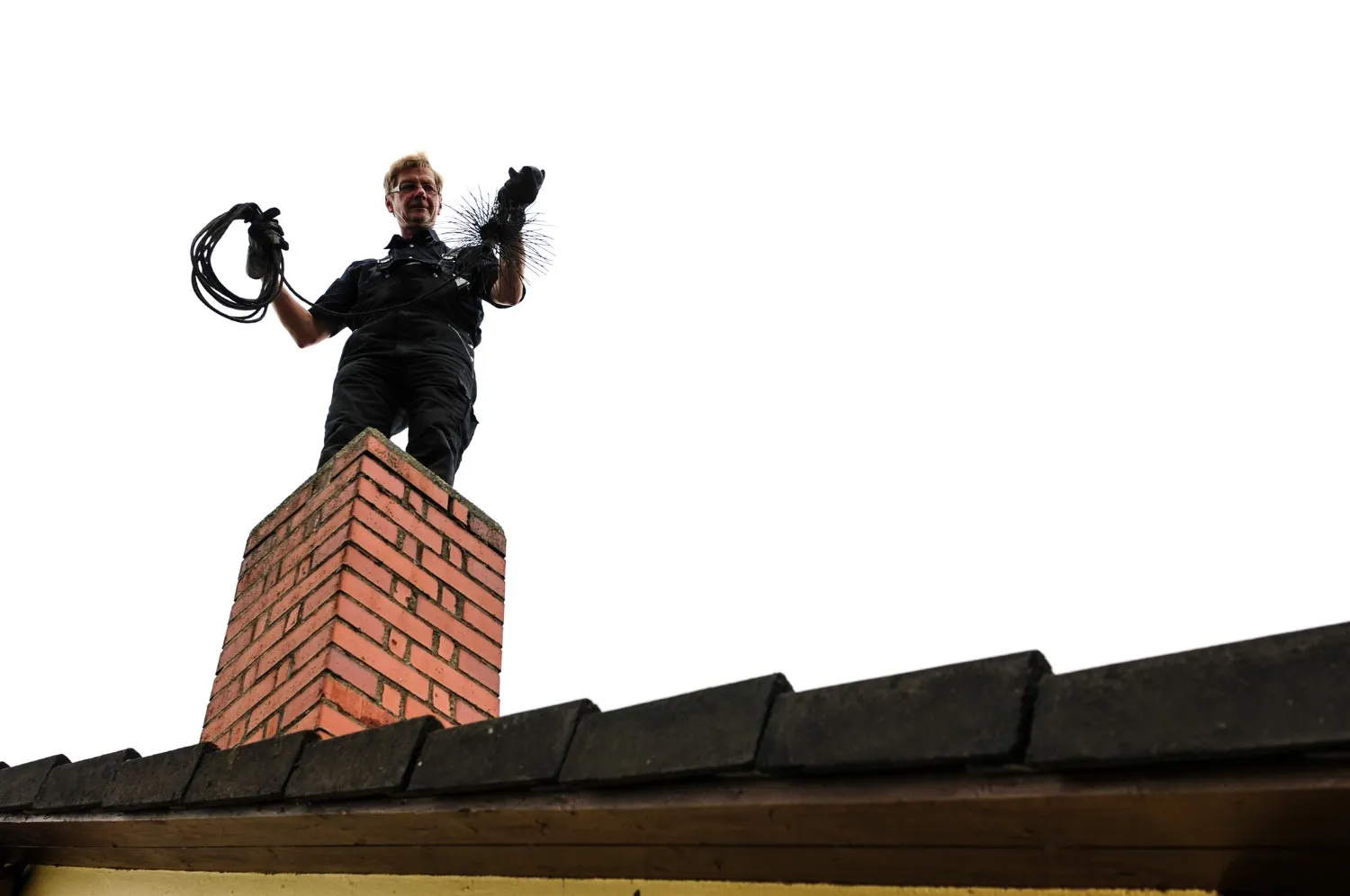 A chimney sweep standing atop a square, red brick chimney stack. The worker, who has light-colored hair, is dressed in a dark uniform and gloves. In one hand, they hold a coil of black rope or cable, and in the other, a large, bristled chimney brush connected to a rod or chain, ready for cleaning the flue. The roof below consists of dark, overlapping tiles. The figure and chimney are sharply silhouetted against an overwhelmingly bright white sky, creating a high-contrast and elevated perspective.
