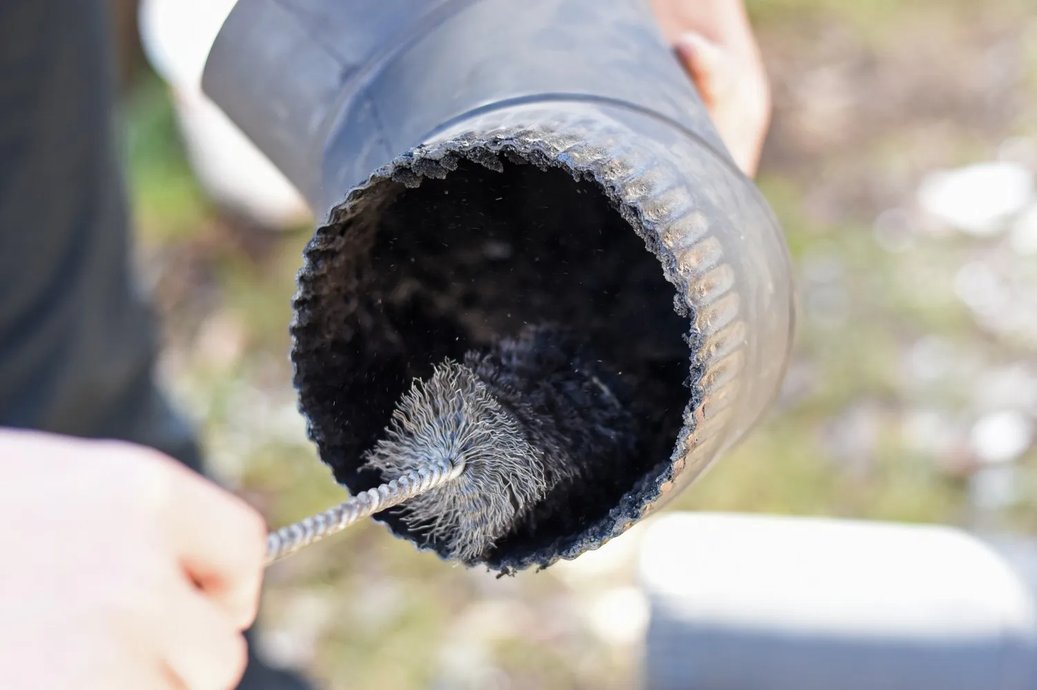 Cleaning of a black metal stovepipe or chimney elbow. A person's hand is holding the pipe while the other hand inserts a small wire chimney brush attached to a coiled metal rod into the opening. The focus is on the brush making contact with the sooty, dark interior of the pipe, which has a slightly rough, serrated edge. The background is a brightly lit, out-of-focus outdoor area.