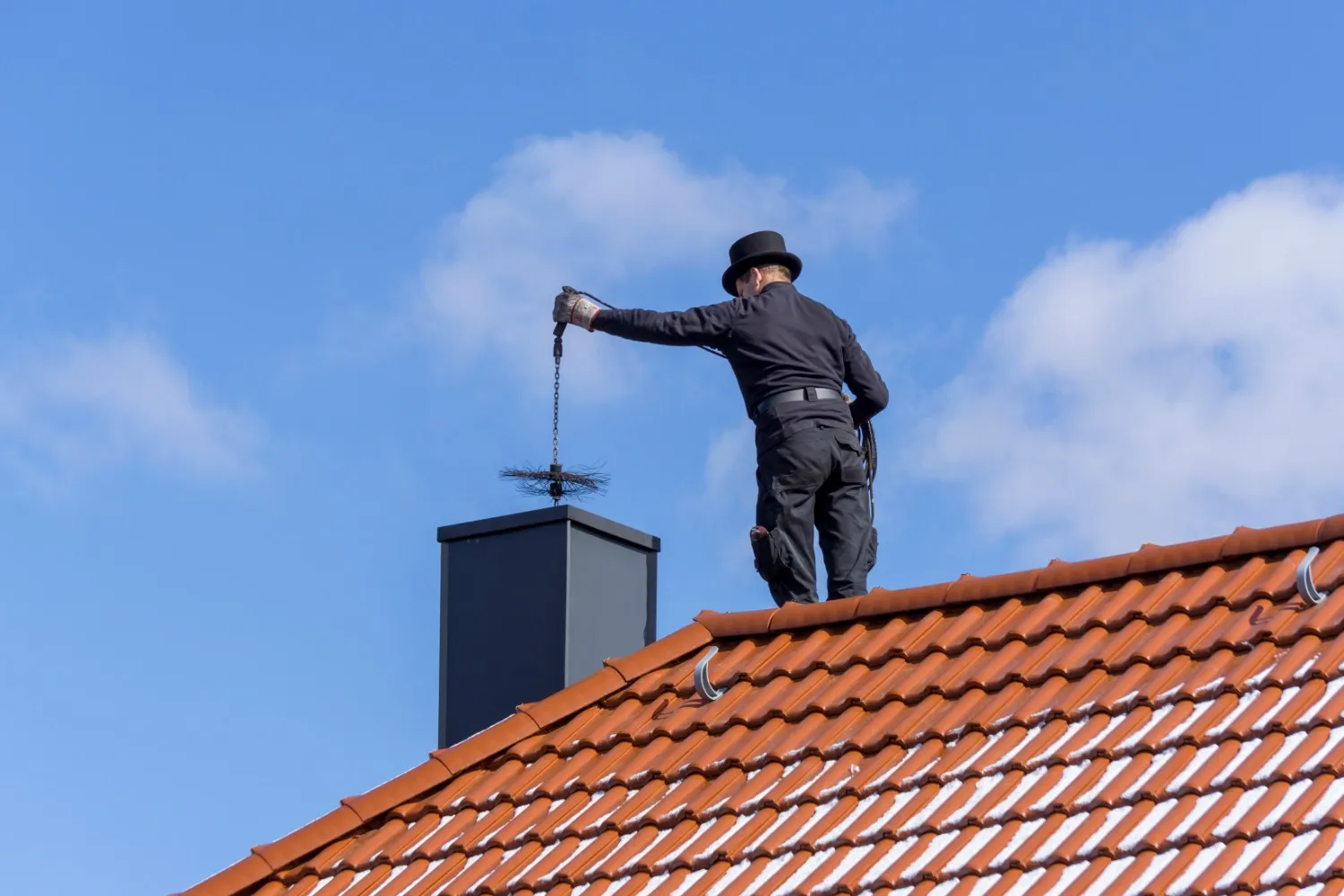 A chimney sweep standing on the orange or terracotta-colored tiled roof of a house against a bright blue sky with scattered white clouds. The sweep is dressed in the traditional black attire, including a top hat and dark work clothes. He is positioned next to a modern, square, dark gray or black metal chimney flue terminal. The sweep is holding a chimney brush attached to a chain, preparing to lower it into the flue opening to begin the cleaning process. The perspective is low-angle, emphasizing the height and the roof's clean, tiled surface, which has some white streaks, possibly from sun reflection or snow.