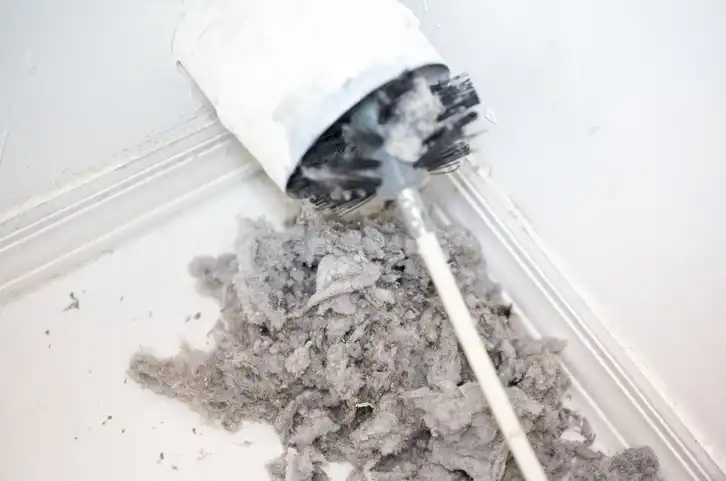 A long-handled brush is being used to remove a large, tangled pile of gray dryer lint from a white vent pipe opening.