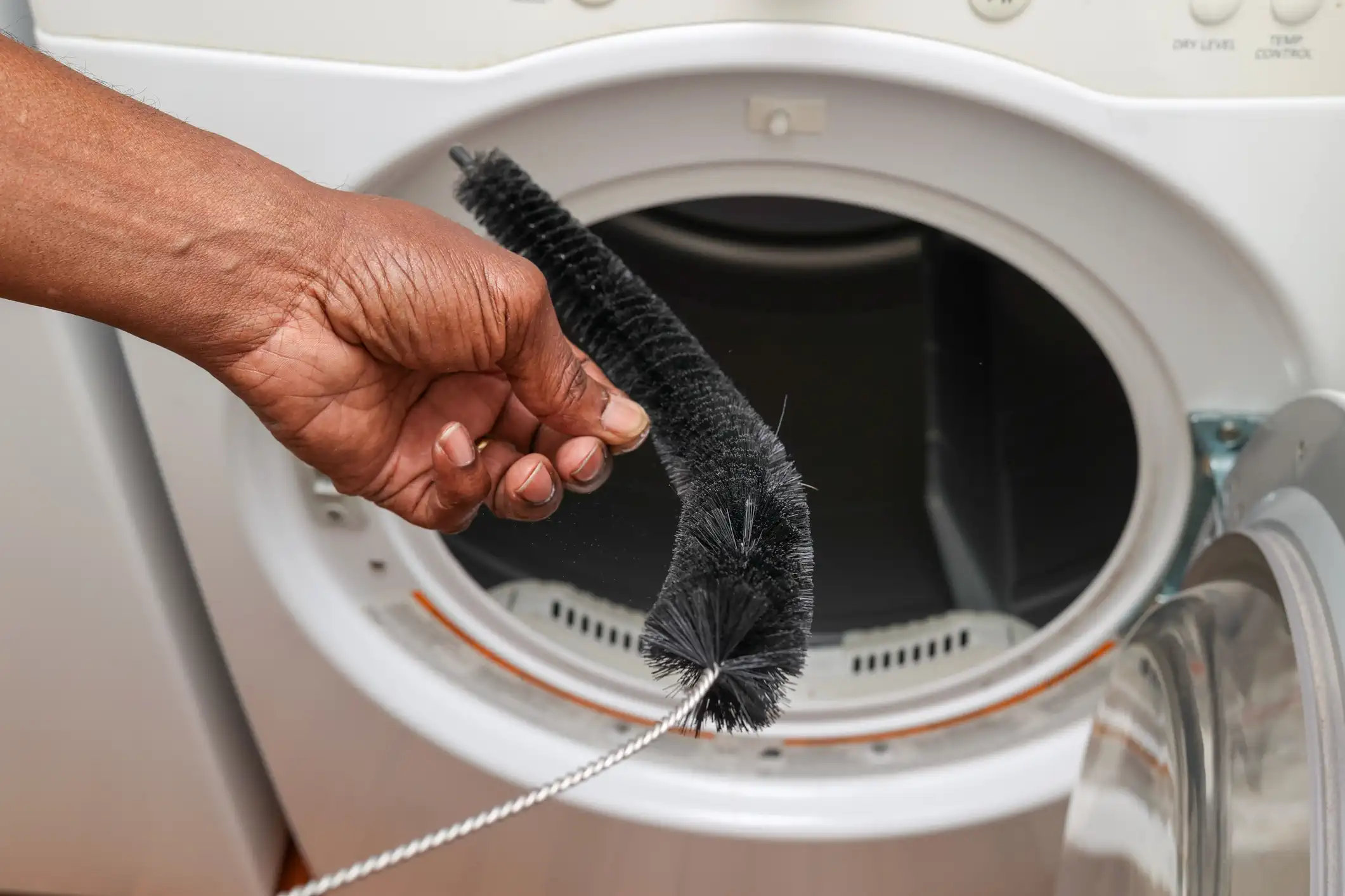 A dark-skinned hand holds a long, flexible dryer vent cleaning brush with stiff black bristles, inserting the end into the open drum of a white front-loading clothes dryer. The image focuses on the cleaning tool and the opening of the dryer where lint and dust collect.