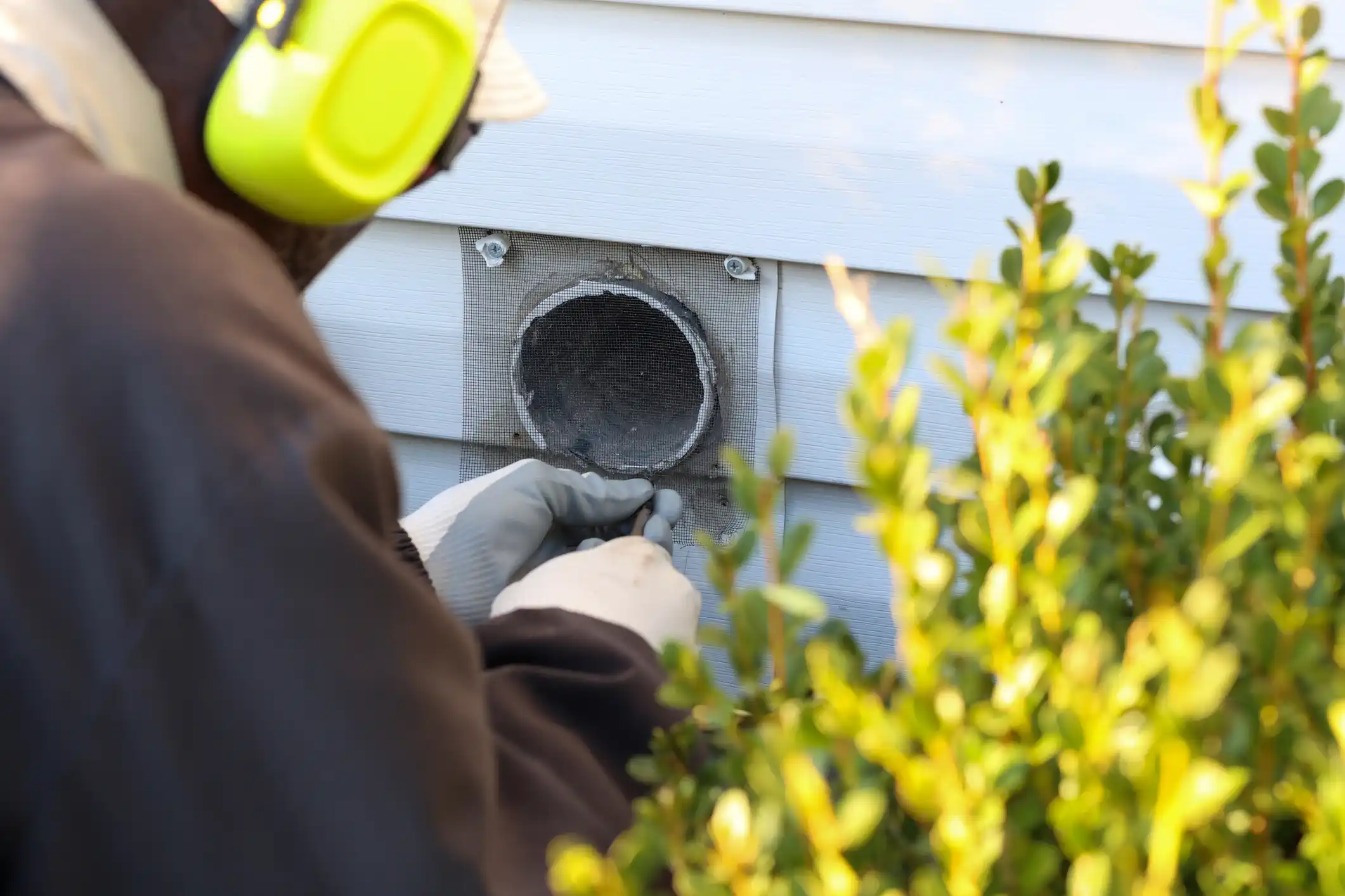 A person wearing a dark jacket, light gloves, and bright yellow earmuffs or hearing protection is cleaning the exterior dryer vent opening on a house. The circular vent is screened and set into light blue siding. The person is using a small tool to clean around the opening, with blurred green shrubbery in the foreground on the right.