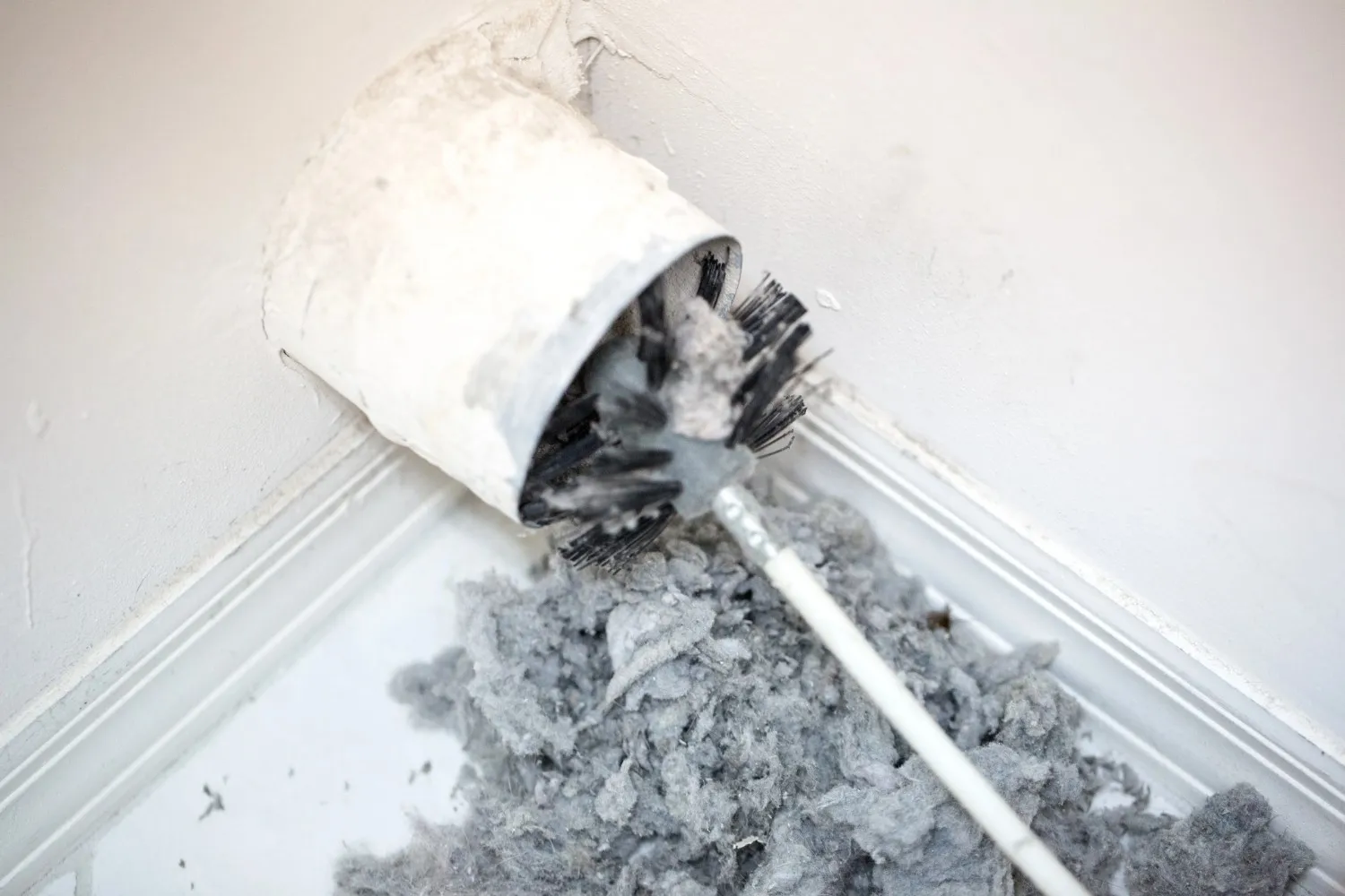 A brush is used to pull a large amount of gray lint and debris from a white dryer vent pipe opening onto a white floor.