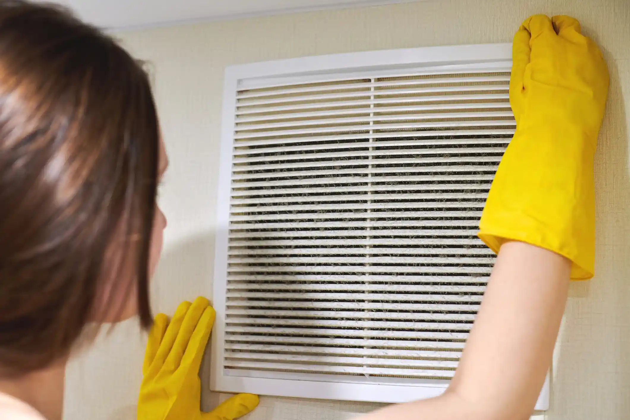 Person in yellow gloves inspecting a wall air vent completely clogged with dust and debris.