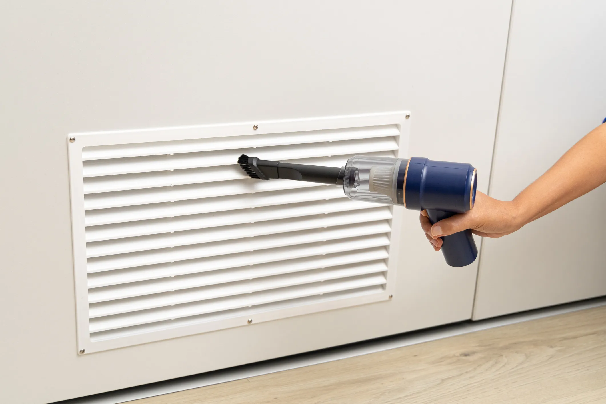Hand using a small handheld vacuum to clean a large white air return grille.