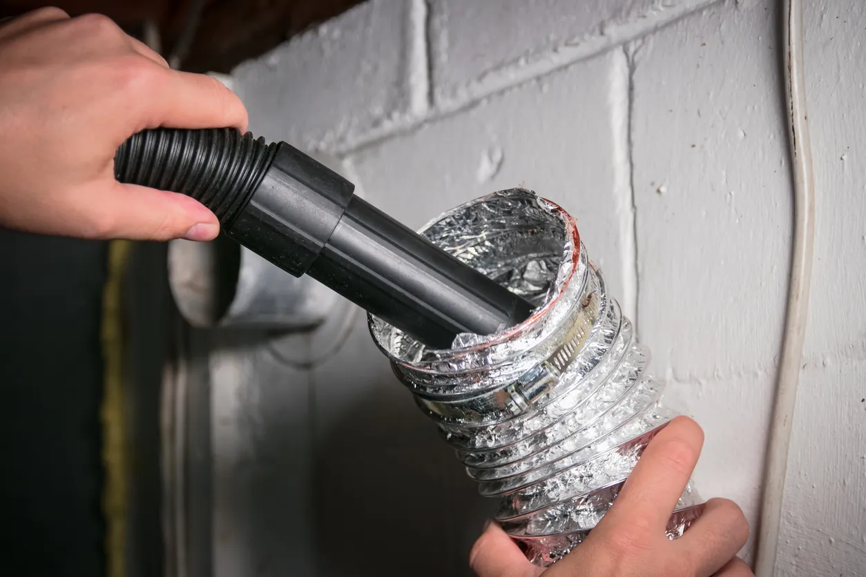 A hand holds a vacuum hose nozzle, inserting it into a flexible metallic air duct to clean the interior.