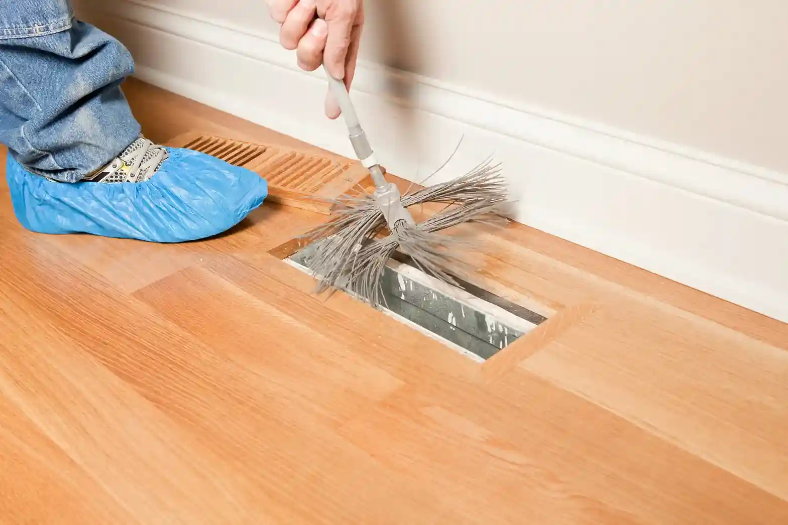 A person wearing blue disposable shoe covers uses a long, flexible brush to clean a dusty floor air vent on a hardwood floor.