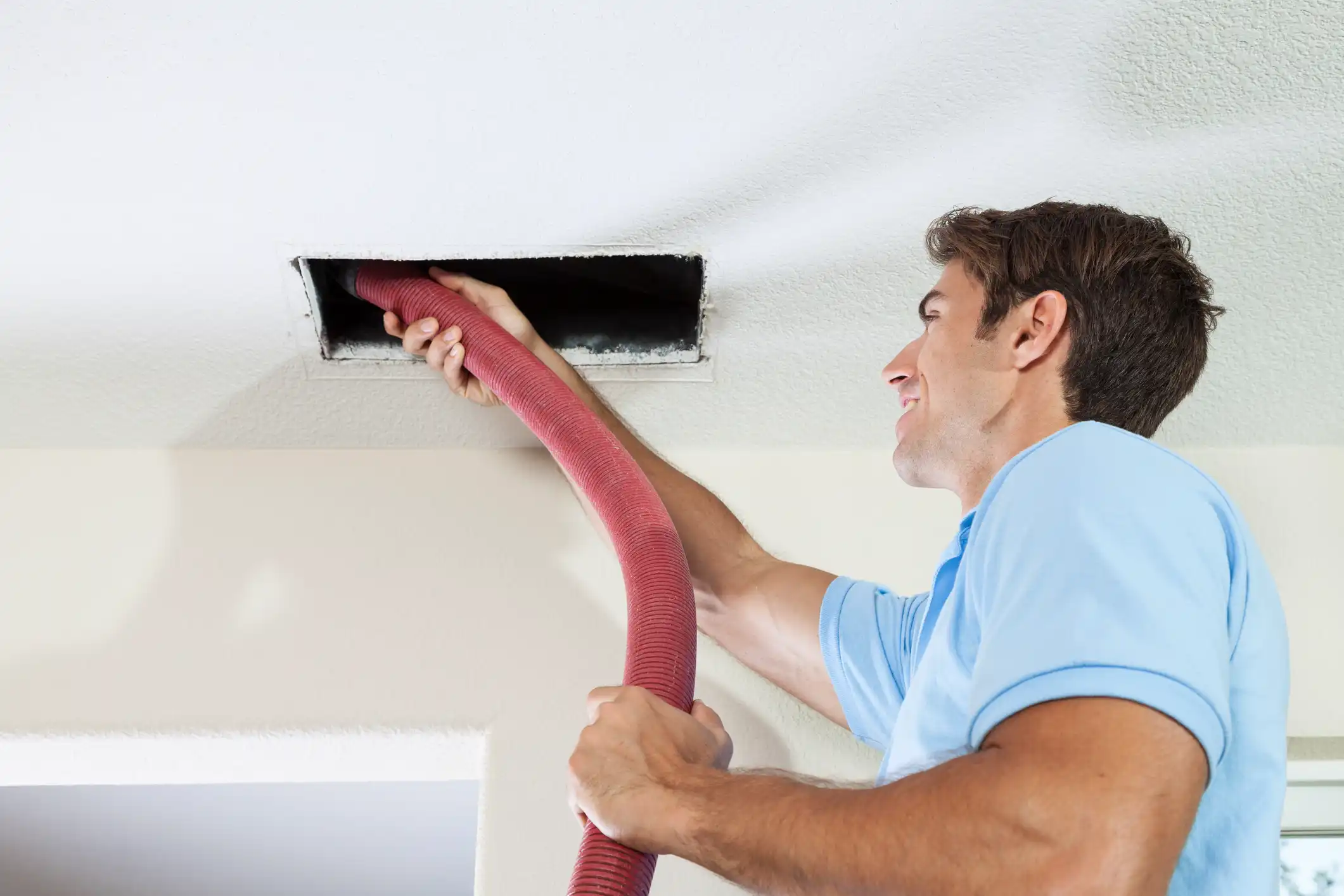 A smiling technician holds a red flexible vacuum hose, inserting it into a rectangular ceiling air vent to clean the duct.