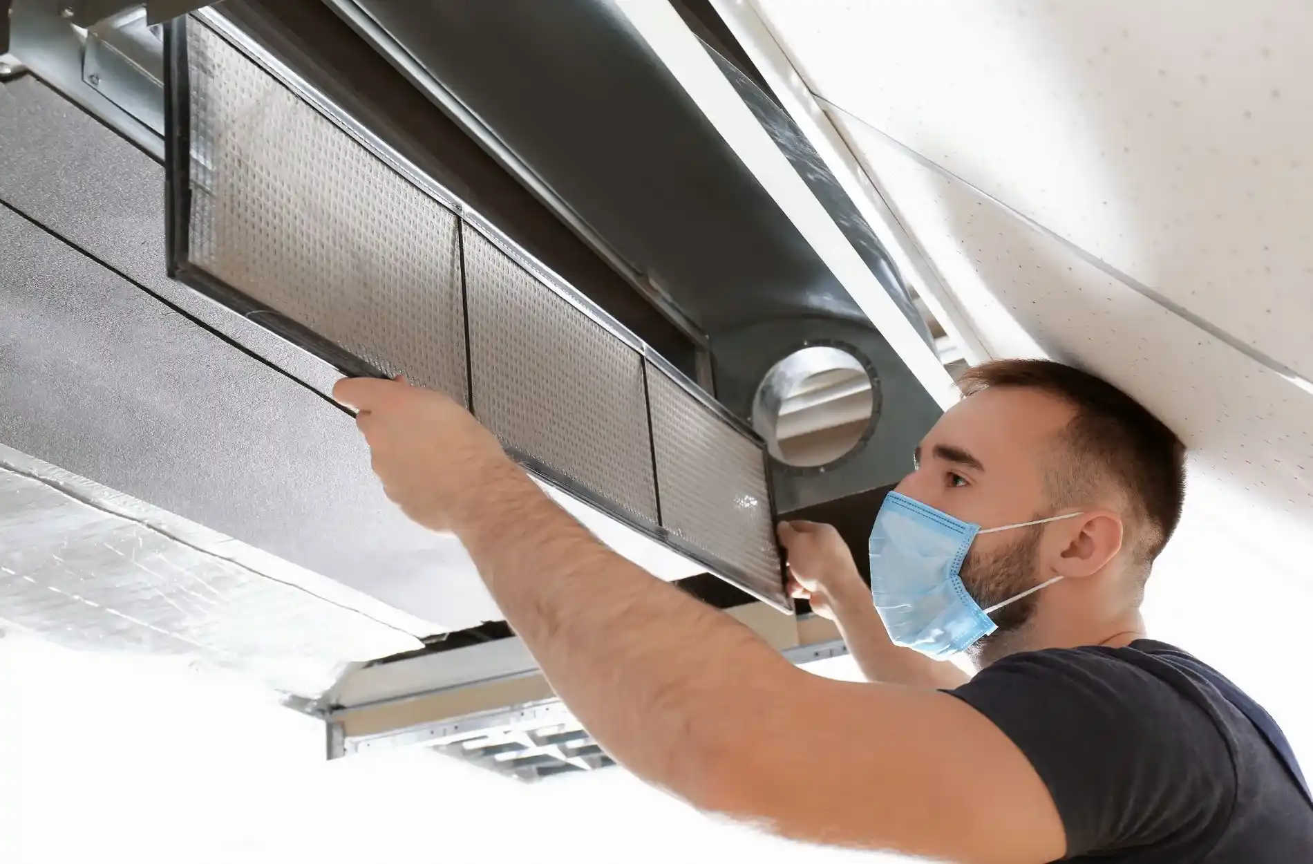 A man wearing a surgical mask installs or removes a rectangular metal air filter from a large suspended commercial air duct system.
