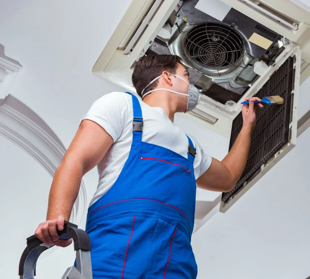 A technician in blue overalls and a mask stands on a ladder, using a small brush to clean a ceiling-mounted HVAC unit's filter.