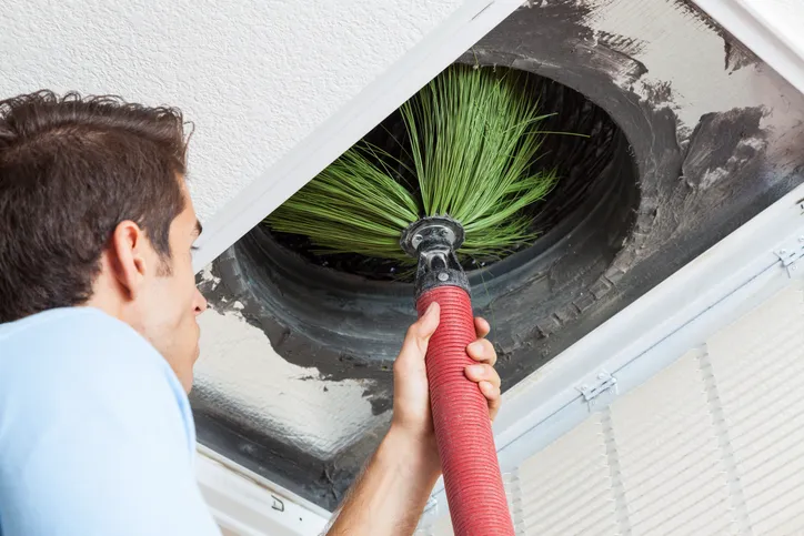 A man uses a round, spinning brush to clean a circular air duct vent in a ceiling.