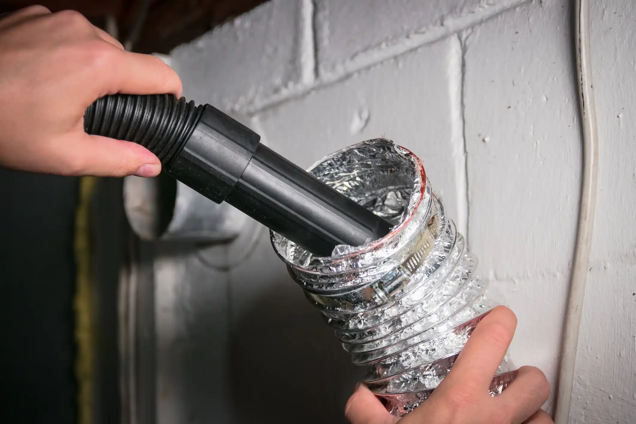 A close-up shot shows a person's hands holding and cleaning a flexible, corrugated aluminum dryer vent or air duct. A black vacuum hose attachment is being inserted into the opening of the duct to remove lint and debris. The duct is secured with a metal clamp and is set against a painted white cinder-block wall in a basement or utility area.