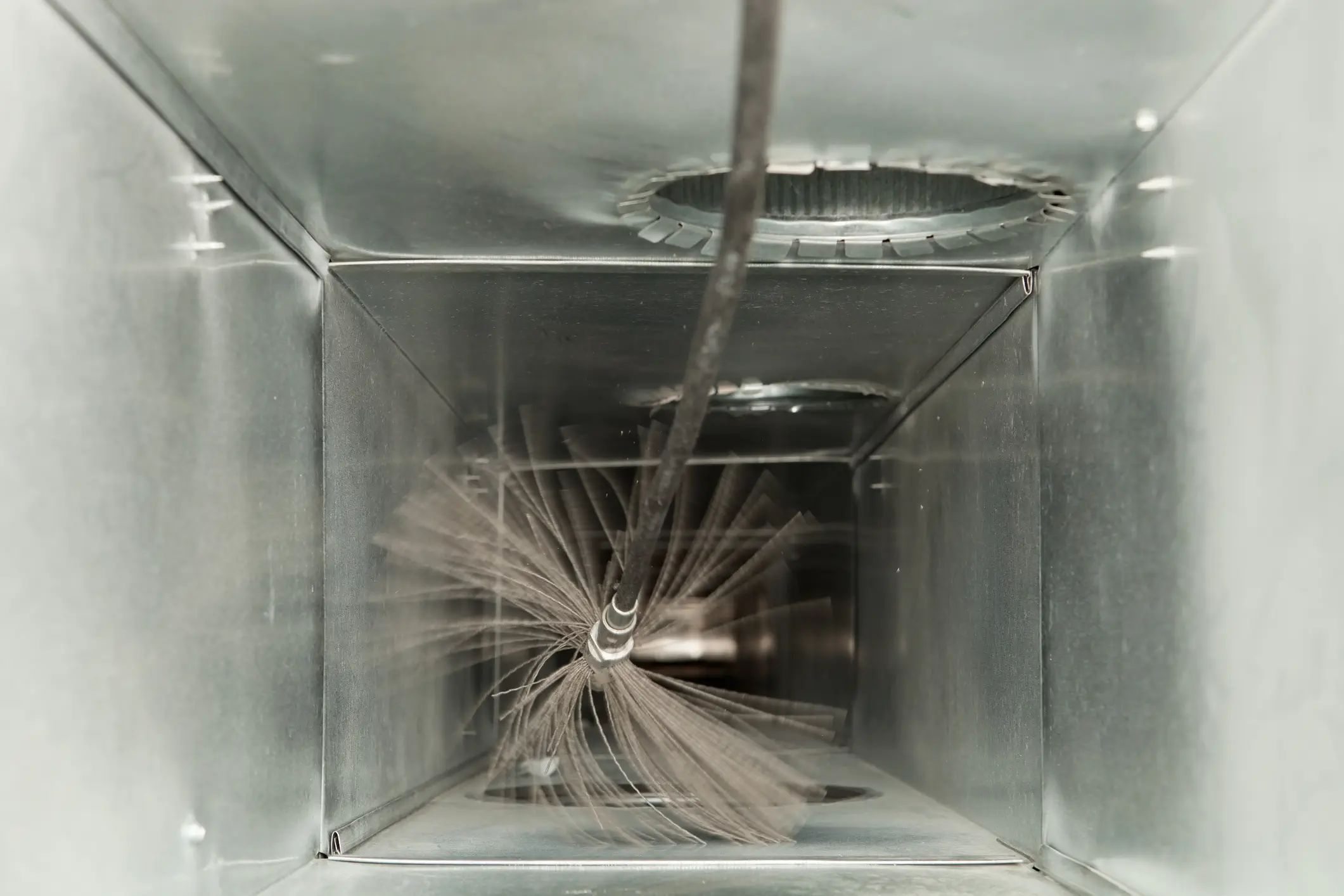 An internal, low-angle view looking down the length of a clean, rectangular galvanized metal duct (plenum or trunk line). A rotating chimney or duct cleaning brush with dark, splayed bristles is visible in the foreground, attached to a long, thin rod. The action of the brush is slightly blurred, indicating it is in motion and actively cleaning the duct walls. In the background, at the top of the duct, there is a circular opening or takeoff with a serrated edge, suggesting a connection point for another duct run. The interior of the duct is highly reflective, showing a metallic surface with some minor dust accumulation.