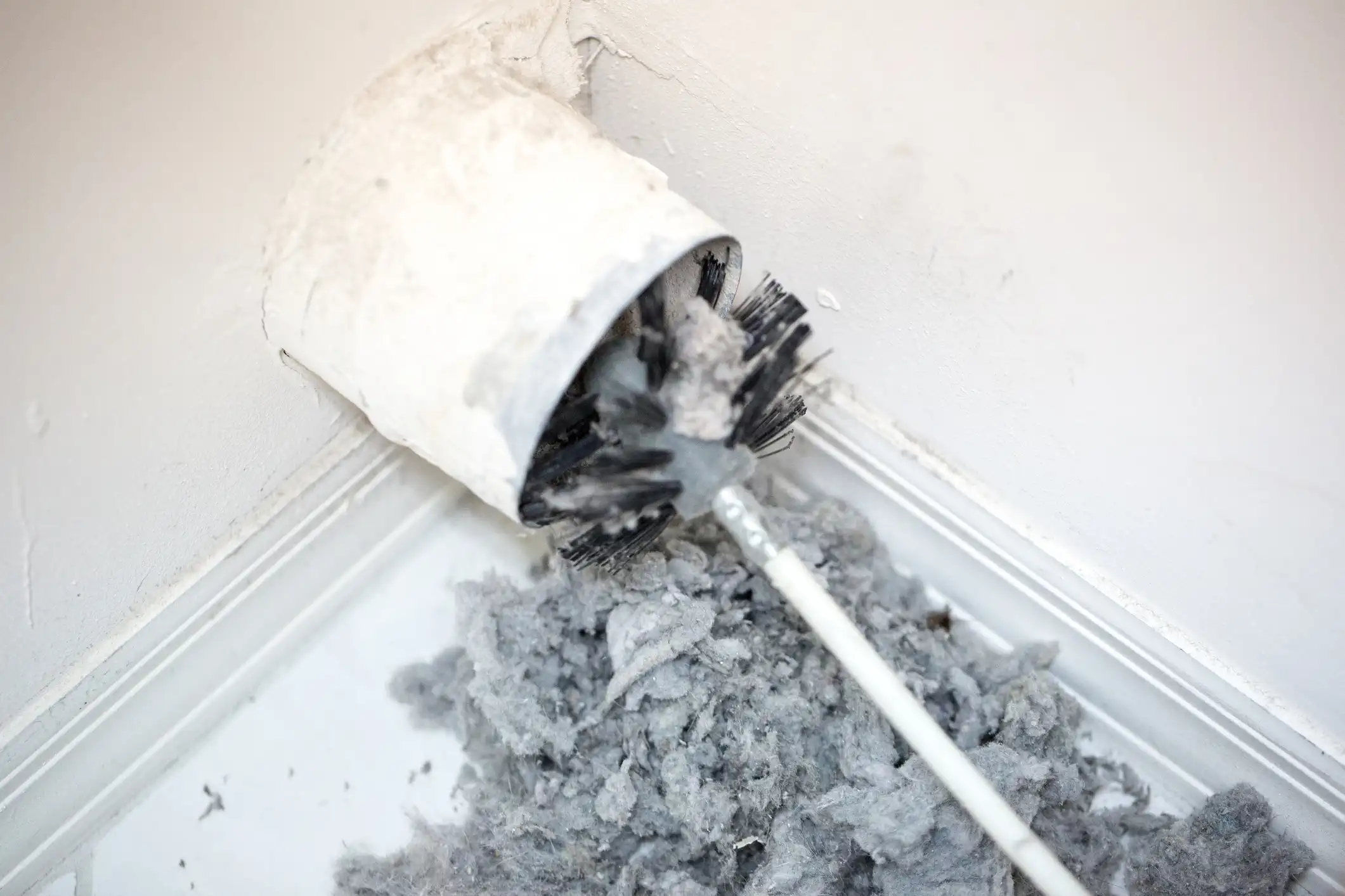 A thick layer of gray lint and dust is piled up on a white windowsill or floor, having been extracted from a white, circular dryer vent or duct. A long-handled brush with black and white bristles, covered in the same debris, is still partially inserted into the duct opening.