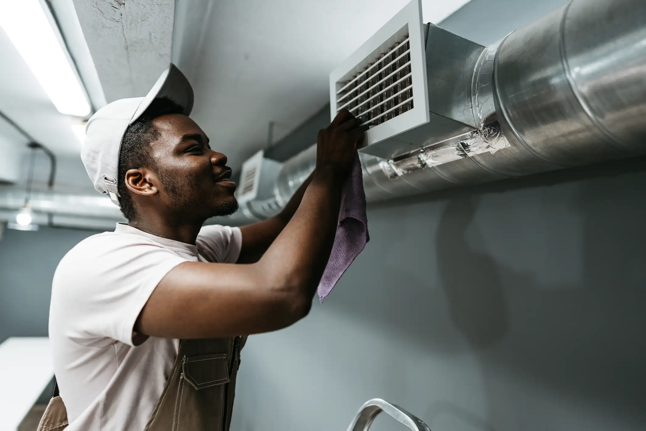 A smiling male technician in a light-colored cap and shirt, wearing what appears to be beige overalls. He is standing on a stepladder (partially visible at the bottom) and is actively working on a rectangular air vent grille mounted on a large, round, galvanized metal duct. He is holding a purple cloth and appears to be cleaning or installing the white grille. The duct runs horizontally along a dark gray wall in what looks like a commercial or industrial space. The overall lighting is bright and overhead.