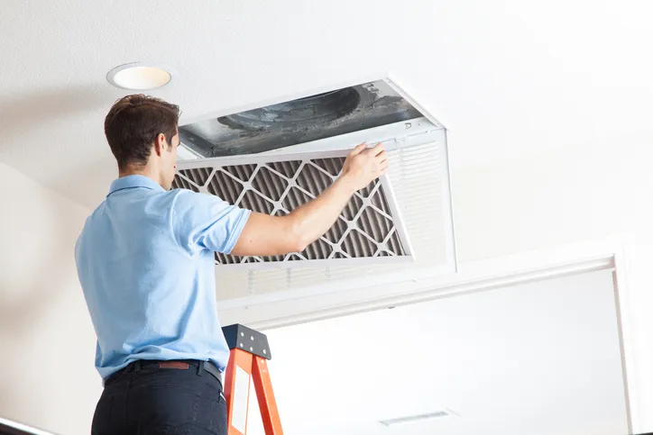 A man on a ladder removes a dusty air filter from a ceiling air duct vent.