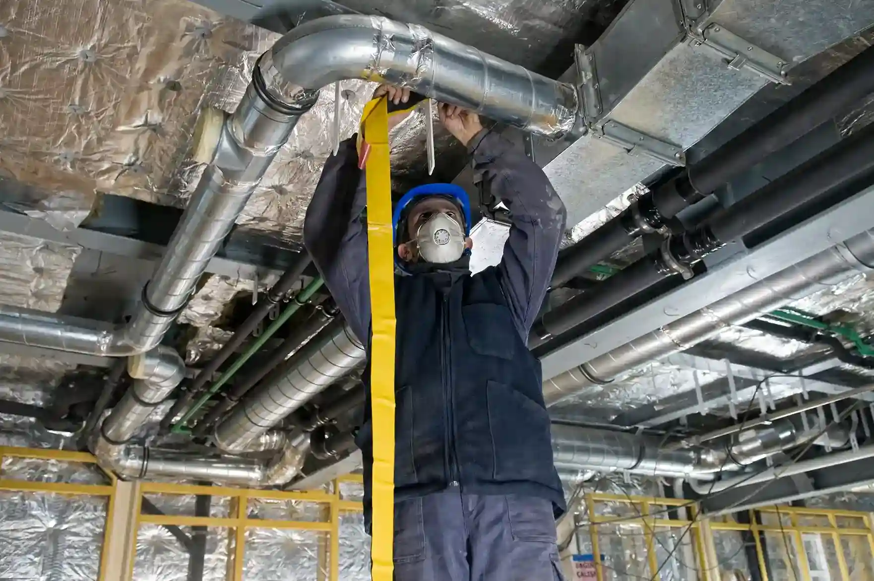 A worker wearing a face mask and hard hat stands on a ladder, using a bright yellow strap to secure an insulated duct in an industrial or commercial space.