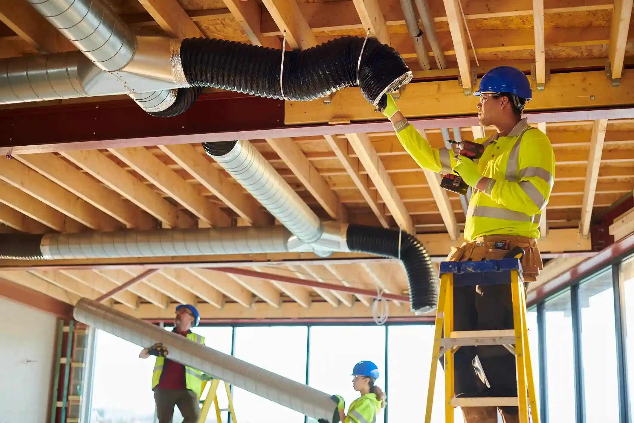Construction workers in hard hats install HVAC ductwork, with one worker on a ladder holding a drill and a flexible duct.