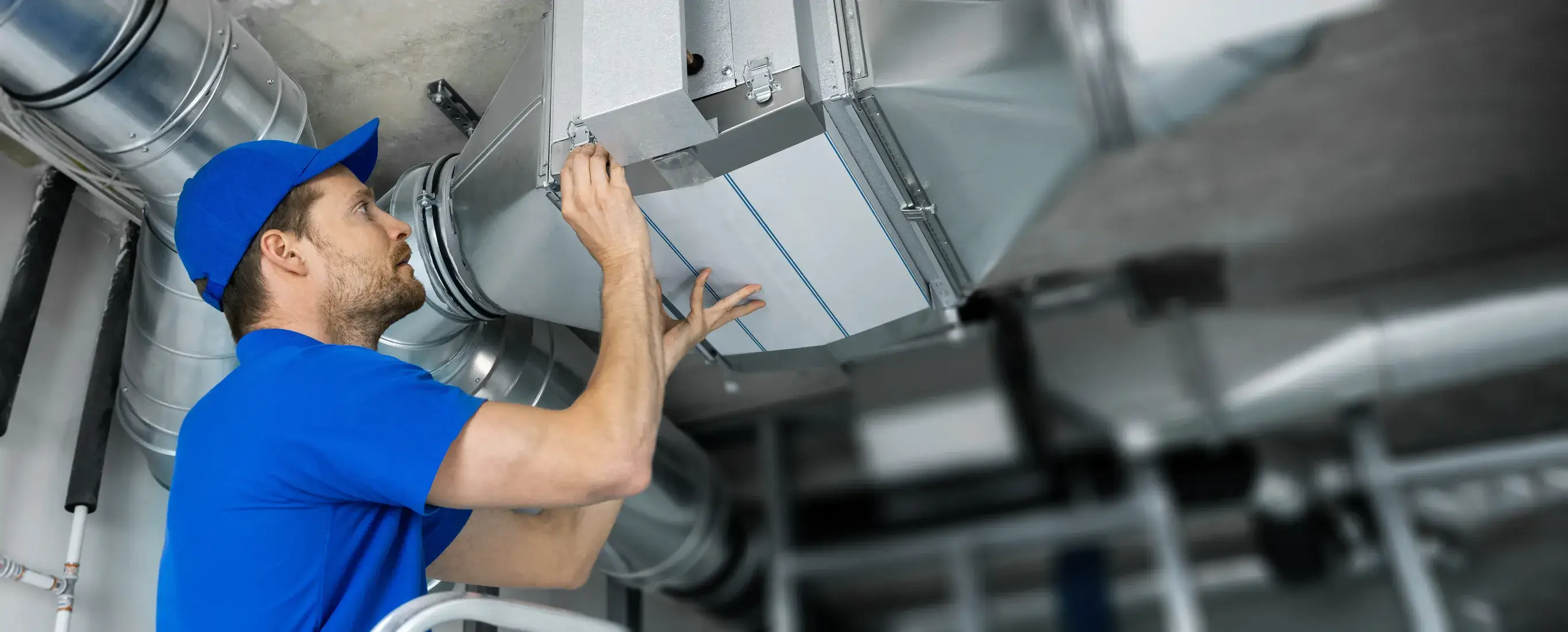 An HVAC technician in a blue shirt and baseball cap smiles while installing or repairing a metal duct system.