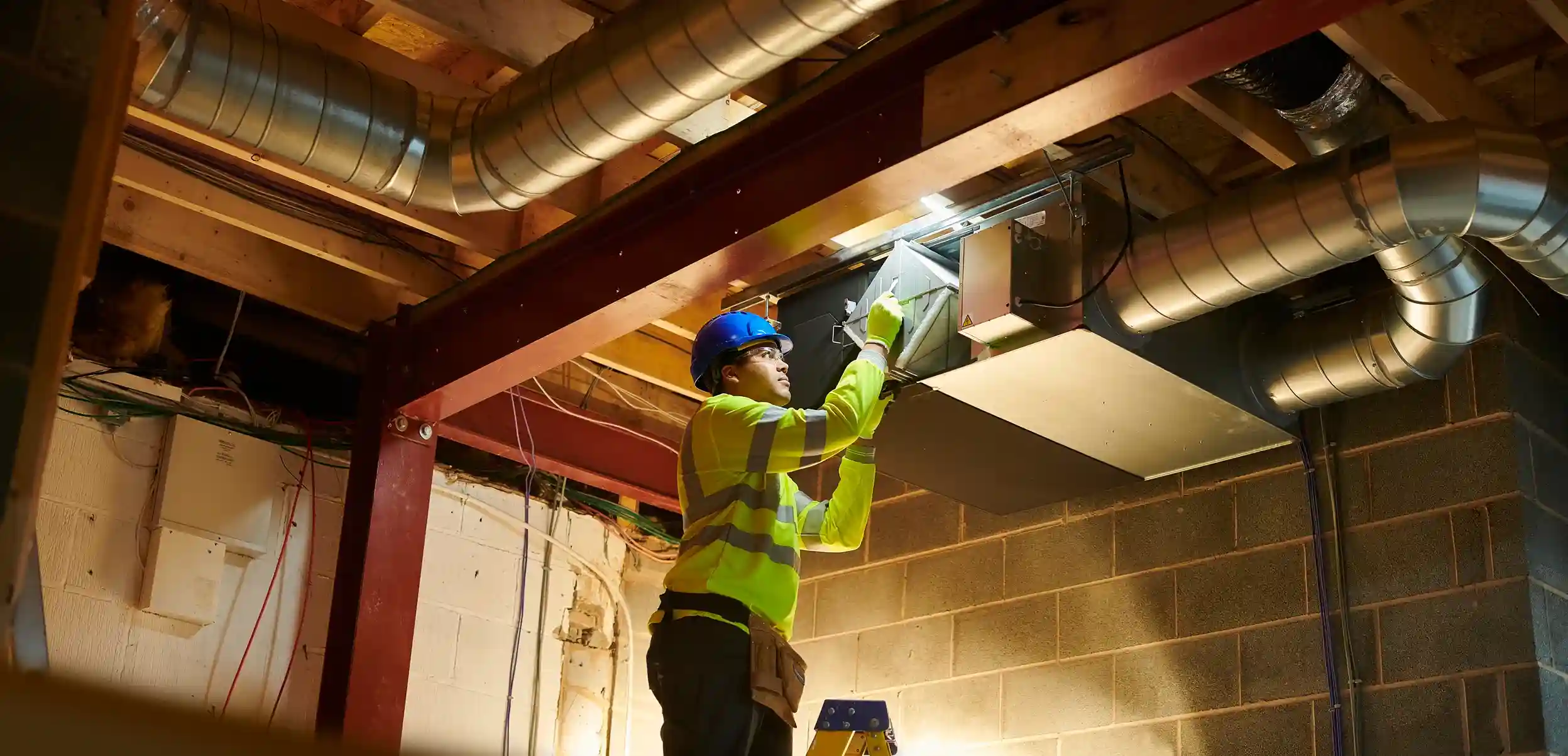 A construction worker in a safety vest and hard hat installs a large rectangular air duct in a basement with exposed wooden joists and a brick wall.