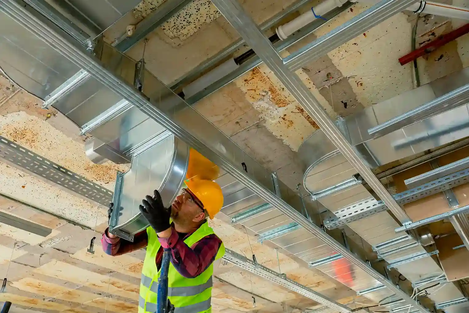An HVAC worker in a hard hat and safety vest holds up a section of a rectangular duct, working on a suspended ceiling grid installation.