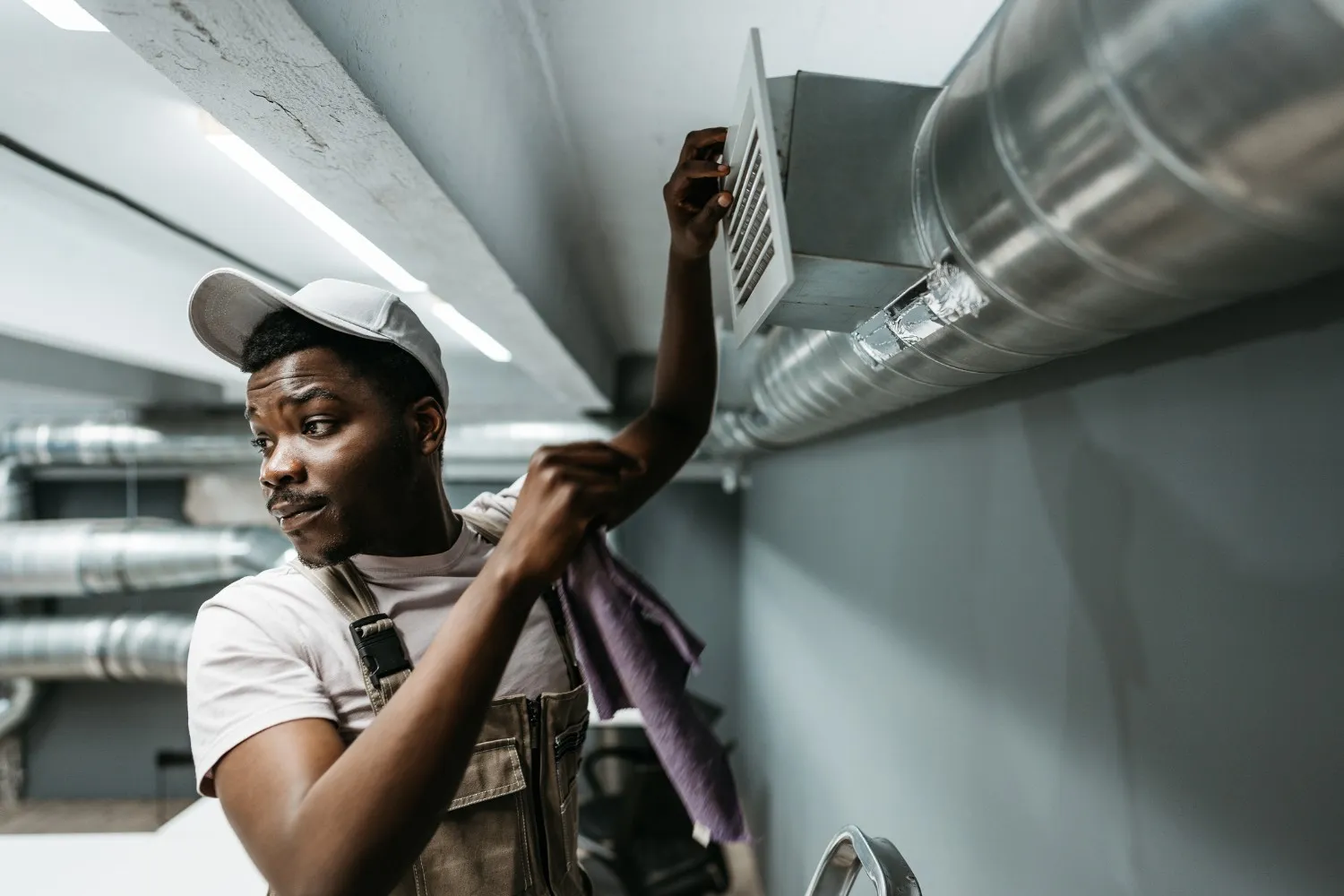  A close-up of a male HVAC technician wearing a white baseball cap and brown overalls. He is carefully removing or adjusting a square white air vent cover attached to a large metal duct on a gray wall. He holds a purple cleaning cloth in his right hand.