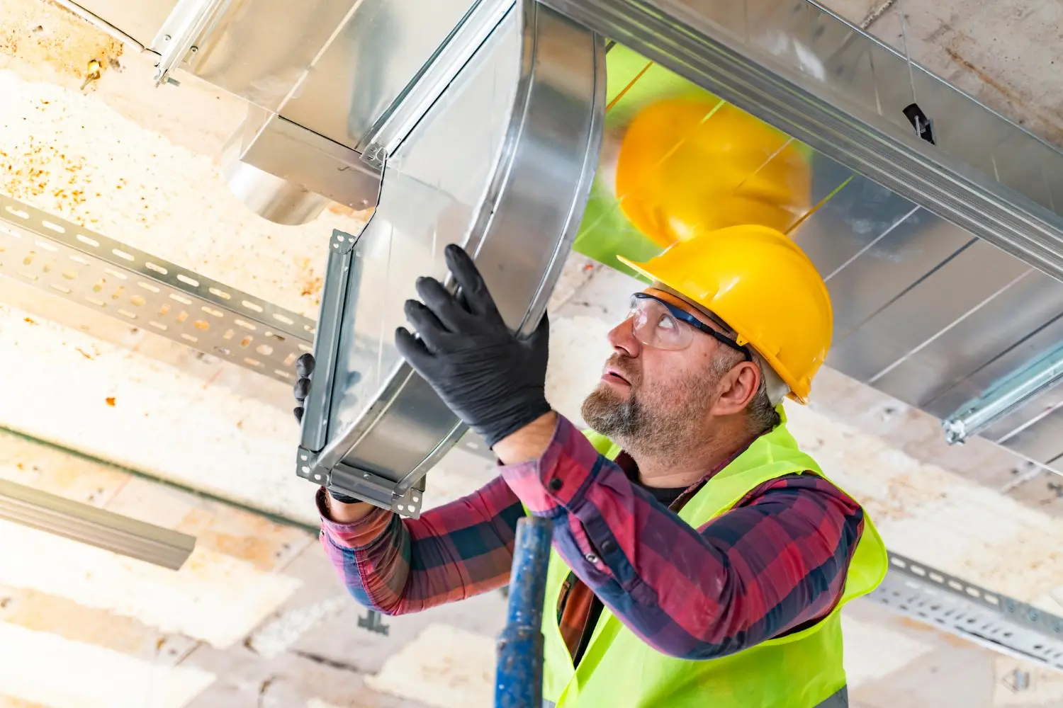 A male HVAC worker in a yellow hard hat, safety glasses, and a high-visibility vest over a plaid shirt is installing or inspecting rectangular galvanized metal ductwork on a construction site ceiling. He wears black gloves and is holding the duct section in place, with sunlight filtering in from the background.