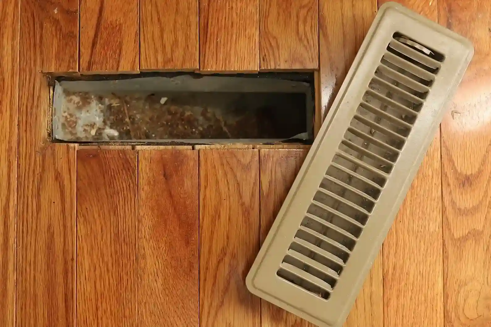 An open floor vent reveals a dusty, dirty air duct, with the vent cover lying next to it on a hardwood floor.